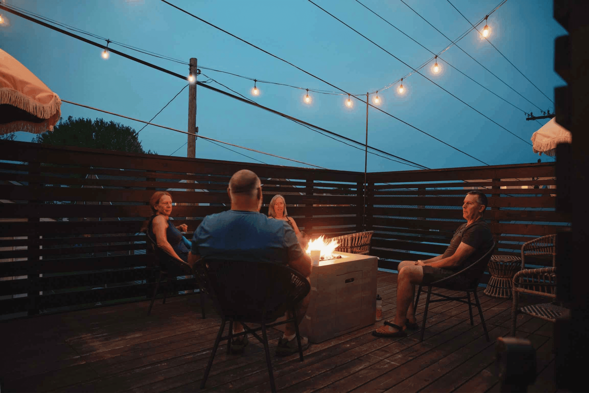 Four people gather around a fire pit on a dimly lit deck, with string lights overhead and a twilight sky.