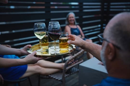 A person serves a tray of drinks, including wine and beer, to a group of people relaxing on a deck.